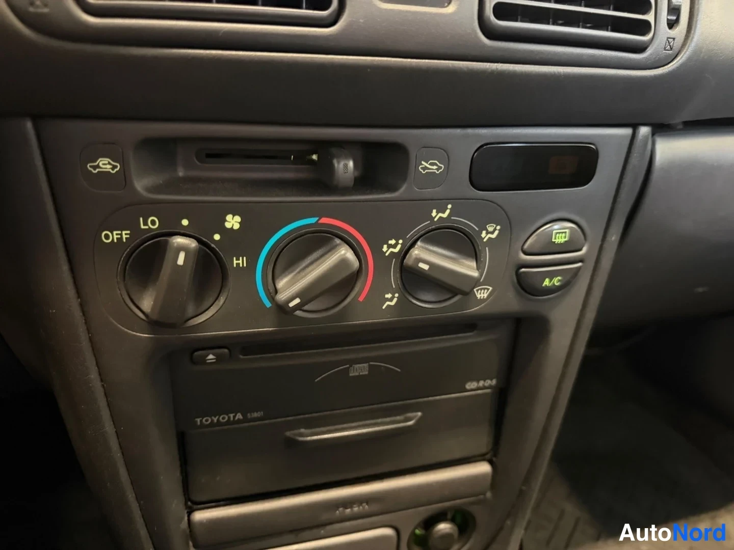 Yksityiskohta: Kojelaudan keskikonsoli: manuaaliset lämmin-/kylmäilmansäätimet ja A/C-painike, alla Toyota-merkitty radio/CD-yksikkö.