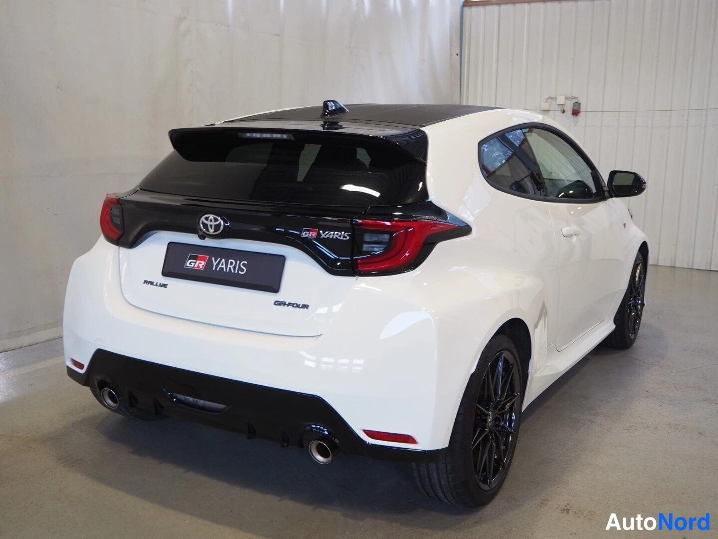 Takaviisto: Valkoinen Toyota GR Yaris takaviistosta sisätiloissa, GR YARIS -merkintä takaluukussa ja kaksoispakoputket.
