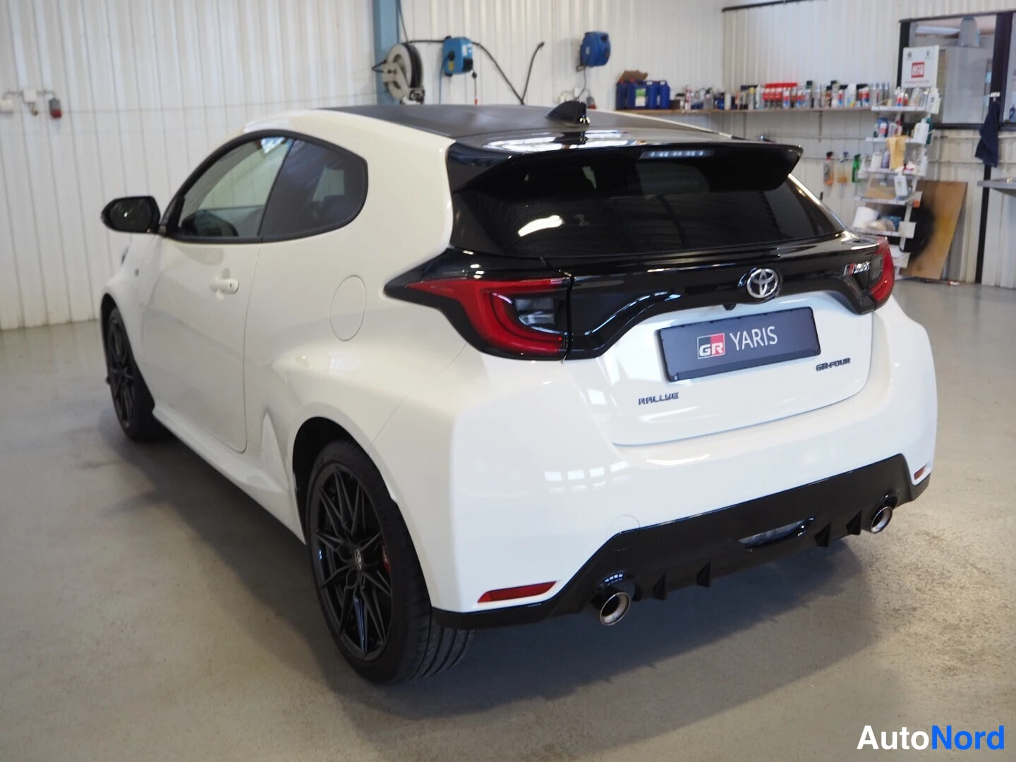 Takaviisto: Valkoinen Toyota GR Yaris takaviistosta sisätiloissa, GR YARIS -merkintä takaluukussa ja kaksoispakoputket.
