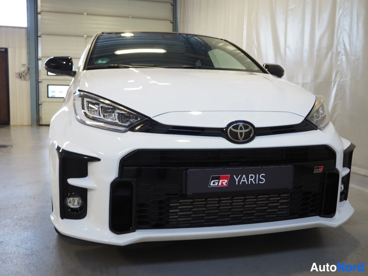 Edestä: Valkoisen Toyota GR Yarisin etupää suoraan edestä kuvattuna, musta etuspoileri ja GR Yaris -kilpi.