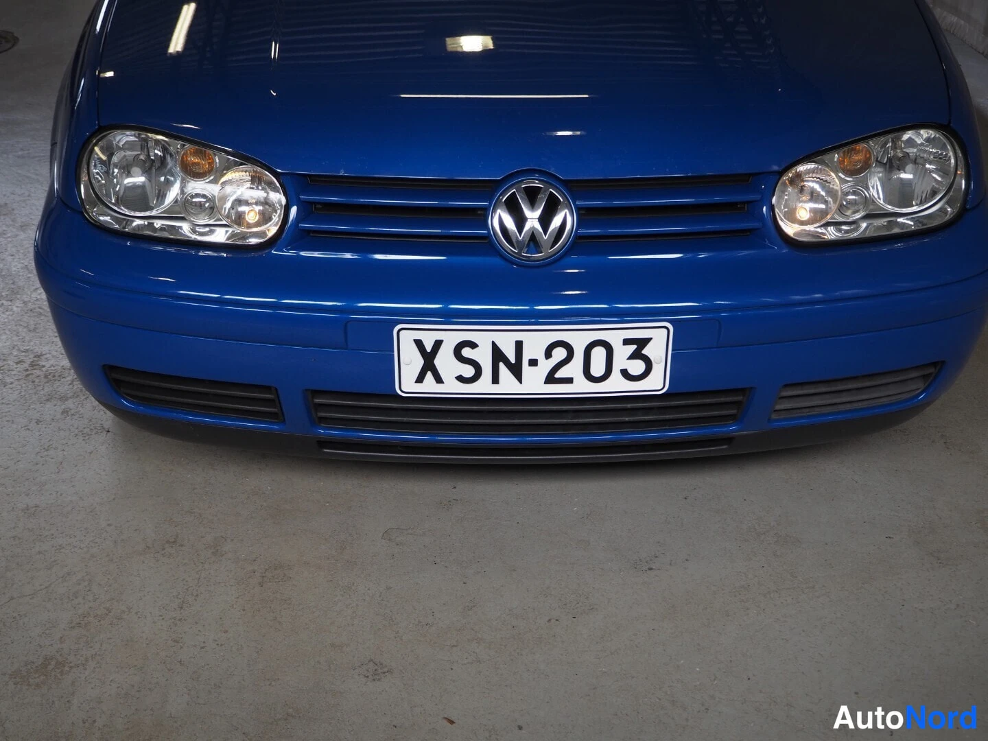 Edestä: Sinisen Volkswagenin etuosan lähikuva: VW-merkki, etupuskuri ja rekisterikilpi.