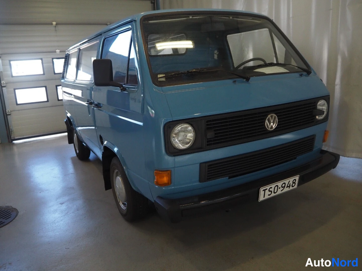 Etuviisto, nokka oikealle: Vaaleansininen Volkswagen Transporter -pakettiauto kuvattu etukulmasta auton oikealta puolella autotallissa, rekisterikilpi.