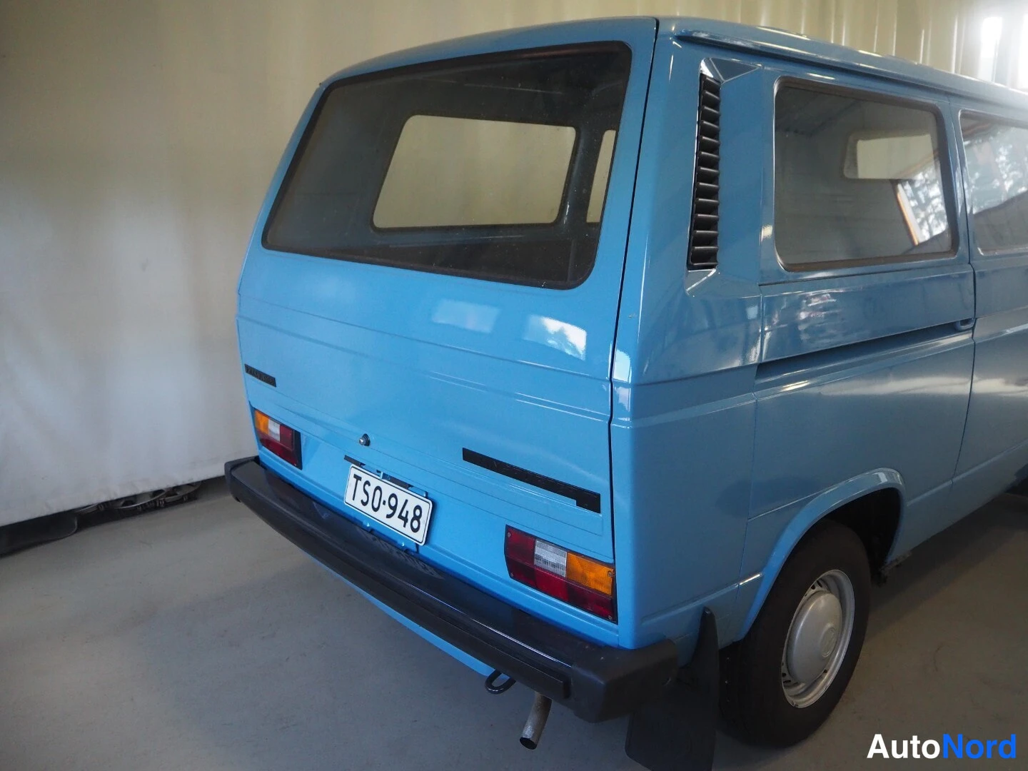 Takaviisto: Vaaleansinisen Volkswagen Transporterin takapää ja kylki kuvattuna takakulmasta, rekisterikilpi ja porrasperä.