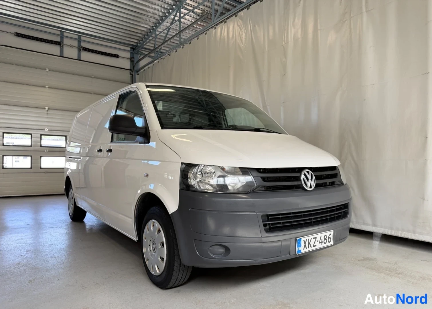 Etuviisto, nokka oikealle: Valkoinen Volkswagen Transporter -pakettiauto kuvattu etukanalta oikealta kulmasta hallitilassa, rekisteritunnus.