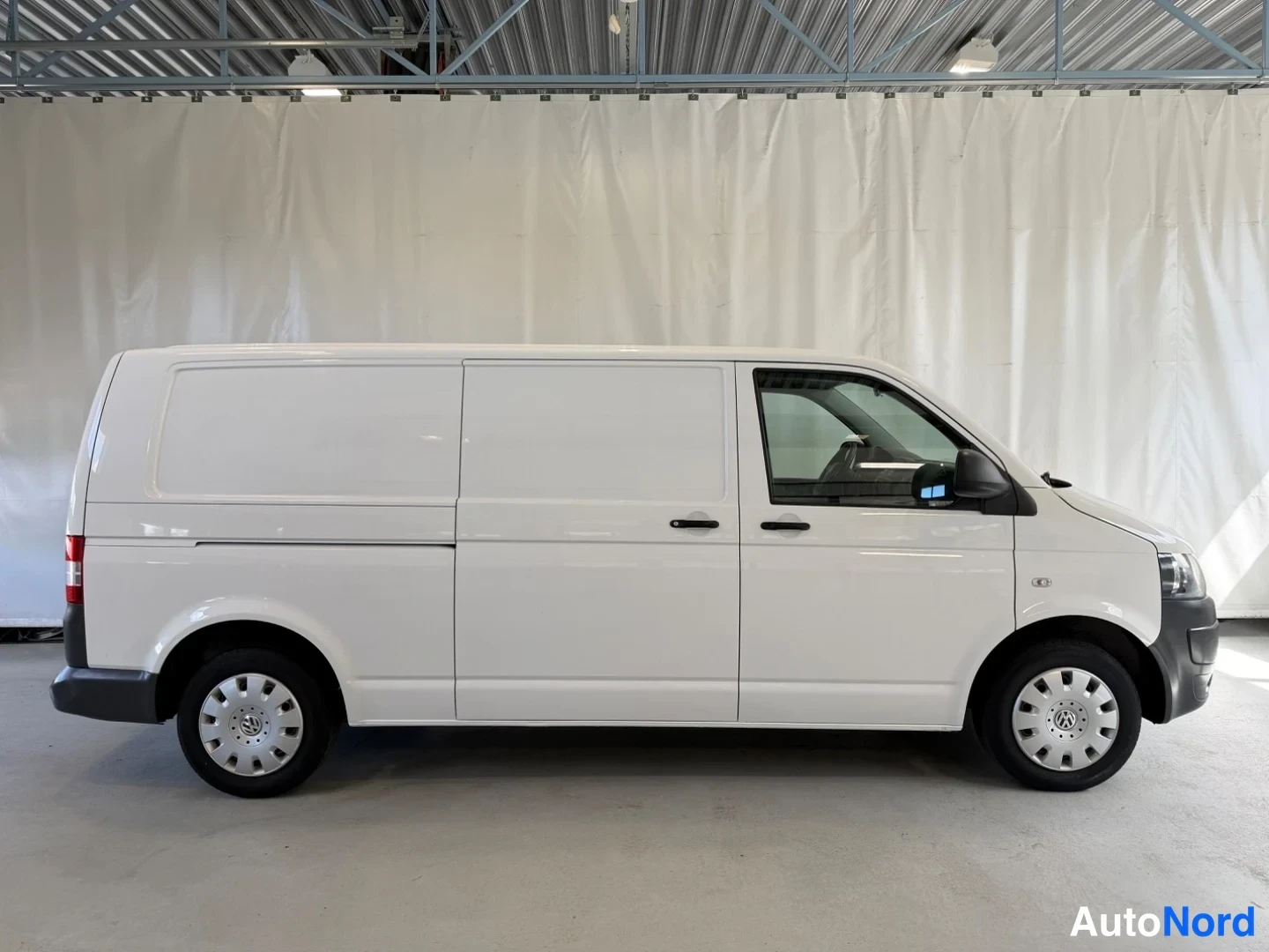 Sivulta: Valkoinen Volkswagen Transporter -pakettiauto sivulta oikealta nähtynä hallissa, liukiovi ja takaovi kiinni.