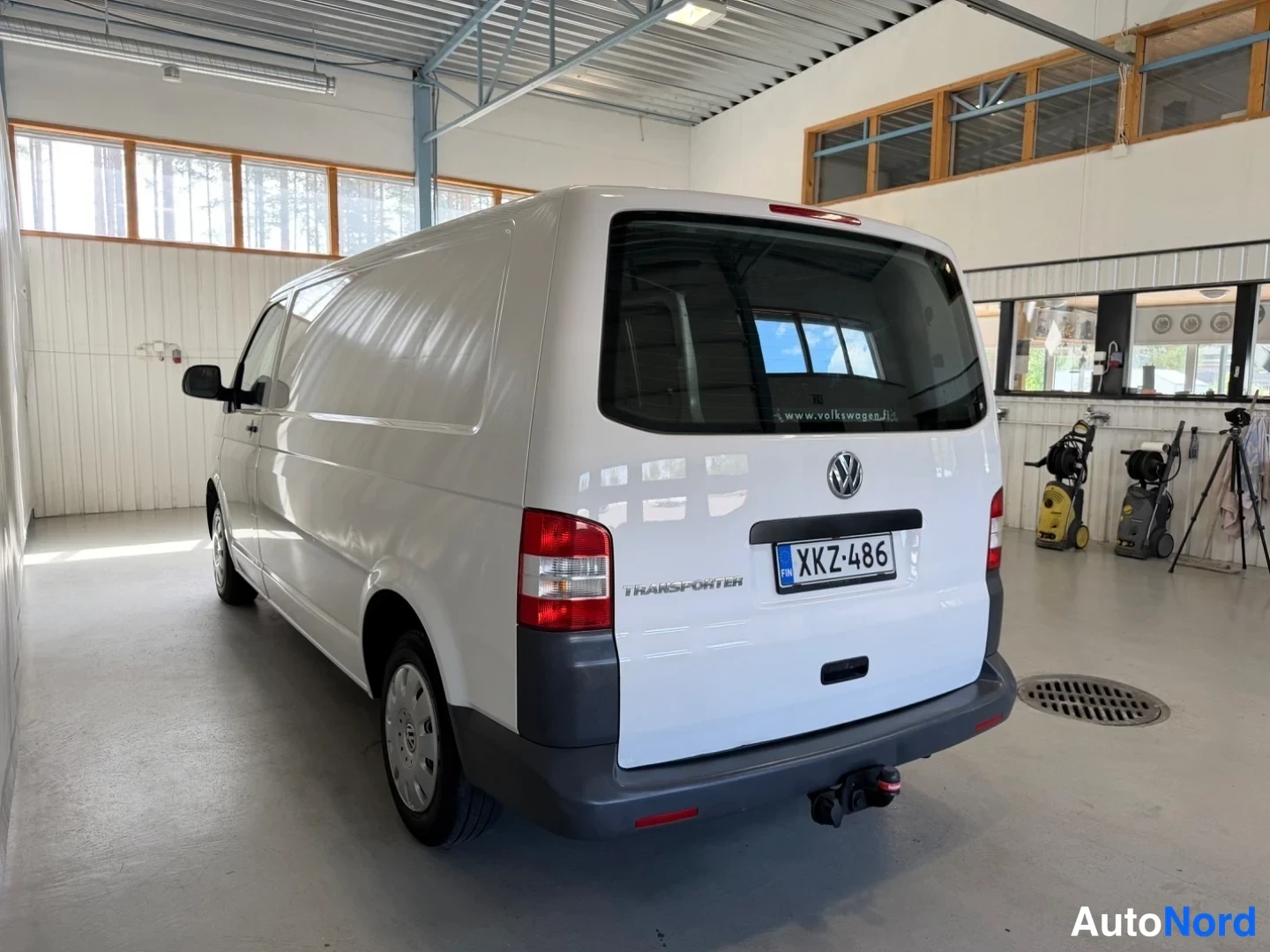 Takaviisto: Valkoinen Volkswagen Transporter -pakettiauto takakulmasta oikealta hallissa, rekisteritunnus ja Transporter-merkintä takaluukussa.