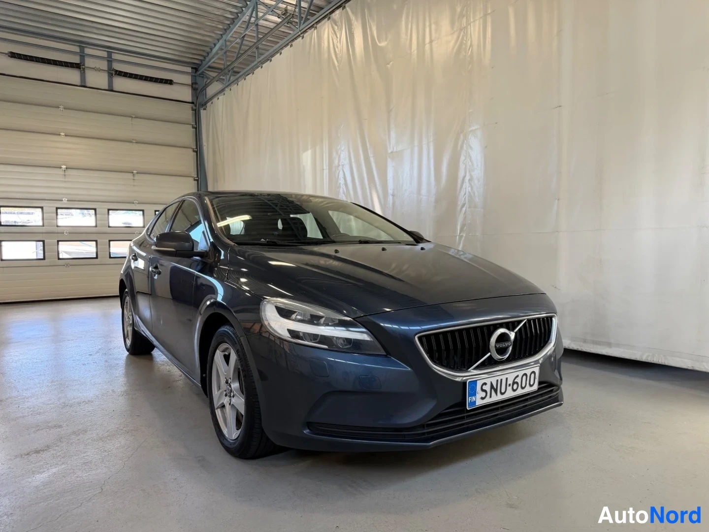 Etuviisto, nokka oikealle: Volvo V40 -henkilöauto tummansinisenä etukulmasta sisätiloissa, rekisterikilpi.
