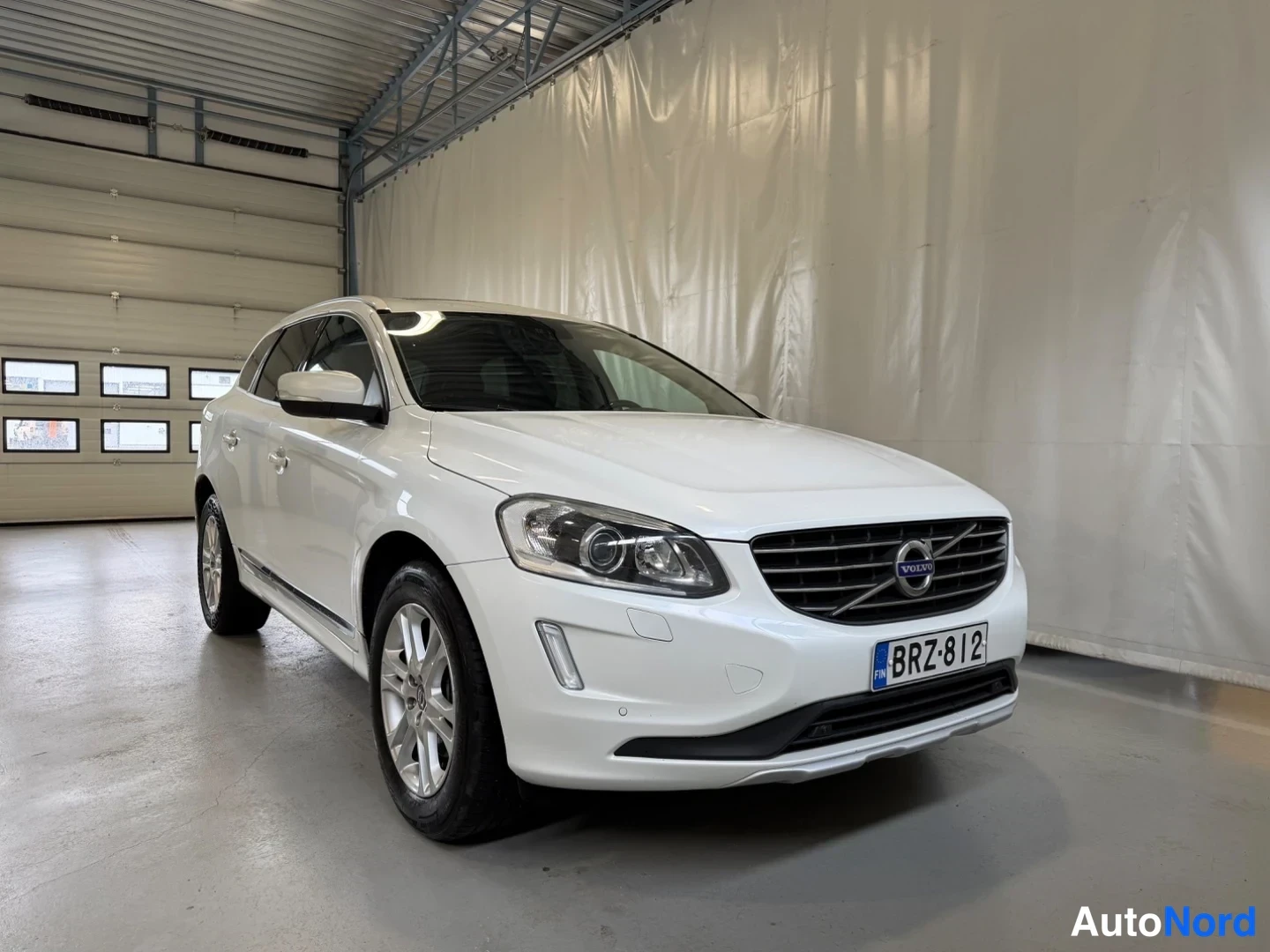 Etuviisto, nokka oikealle: Valkoinen Volvo XC60 näkyvä etuosa ja vasen kylki kolmessa neljäsosassa sisätiloissa, rekisterikilpi.