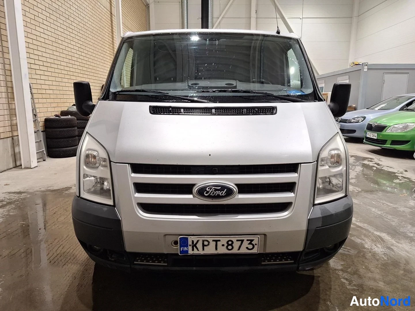 Edestä: Hopeanvärinen Ford Transit -pakettiauto suoraan edestä; etusäleikössä Ford-merkki ja rekisterikilpi, ajovalot ja tumma etupuskuri näkyvät.