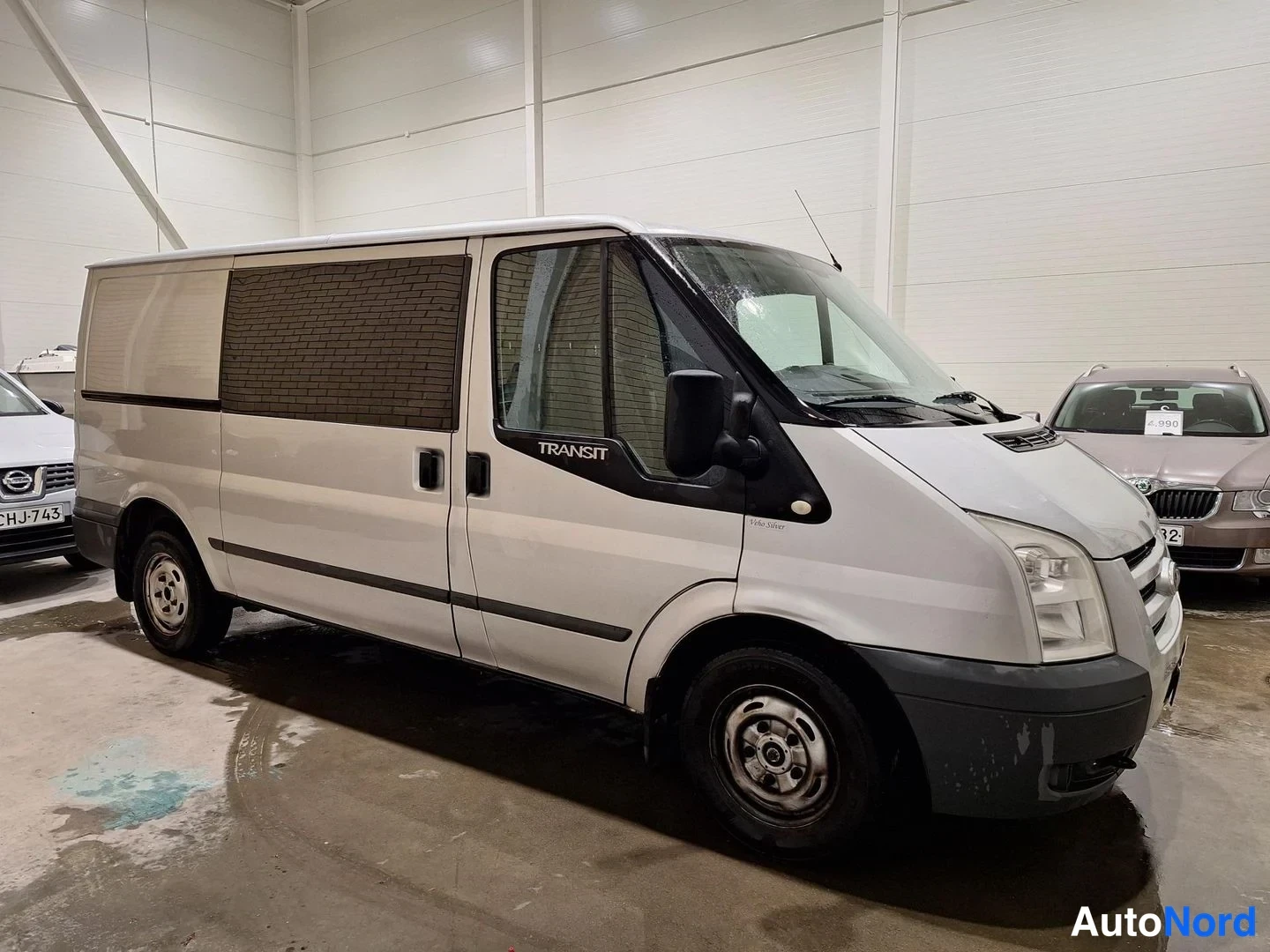Etuviisto, nokka oikealle: Hopeanvärinen Ford Transit -pakettiauto kolmivinkeltyä edestä oikealta sisätilassa; ajovalot, etusäleikkö ja oikea kylki näkyvät, ovessa lukee TRANSIT.