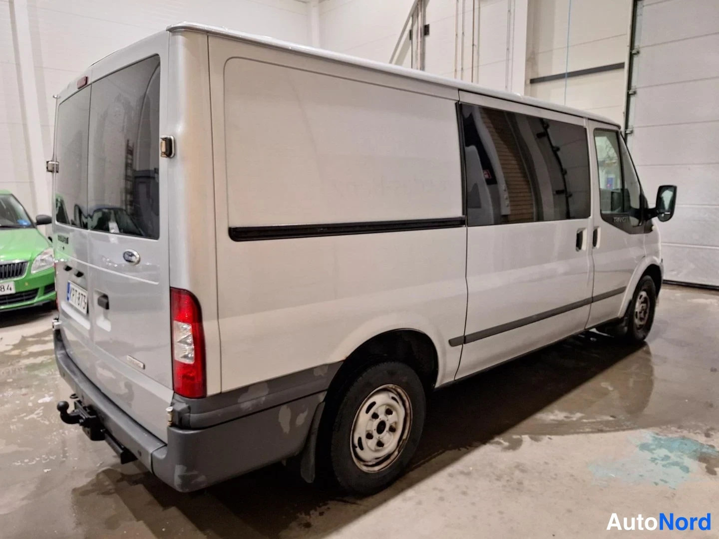 Takaviisto: Hopeanvärinen Ford Transit -pakettiauto kolmivinkeltyä takaa vasemmalta; takavalot, takanosto-ovet, rekisterikilpi ja vetokoukku näkyvät.