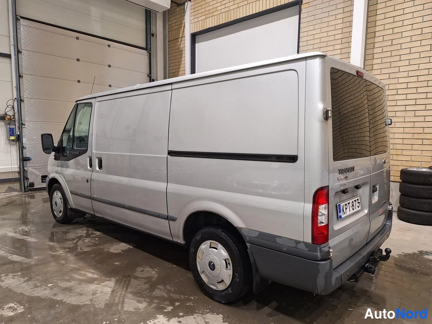 Takaviisto: Hopea Ford Transit -pakettiauto sivulta takaa nähtynä sisätiloissa, takaluukun B-ruuvit auki ja rekisterikilpi.