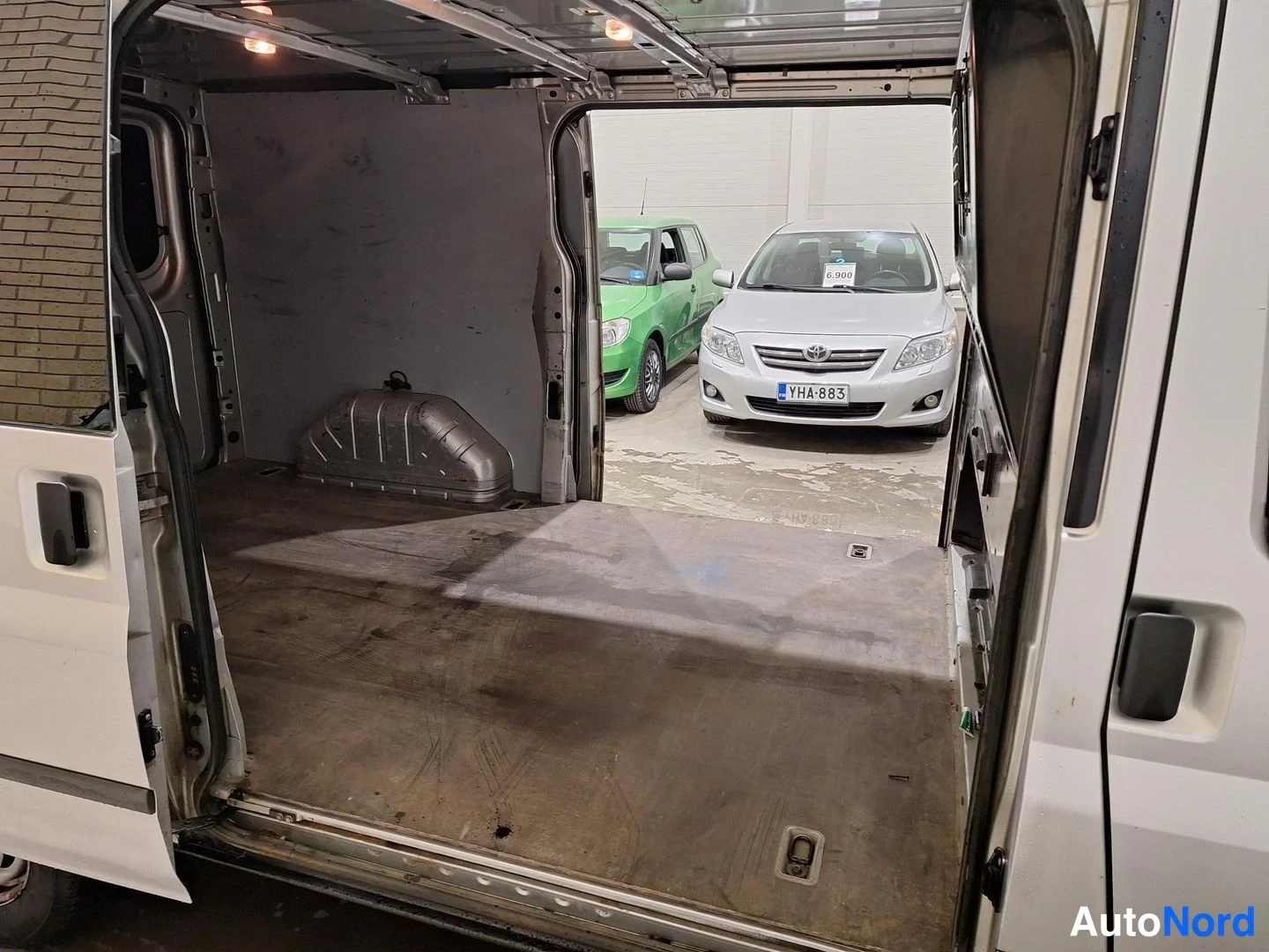 Muu: Avoin pakettiauton tavaratila tyhjine lattioineen ja varallisineen; oviaukon takana autohallissa kaksi henkilöautoa.