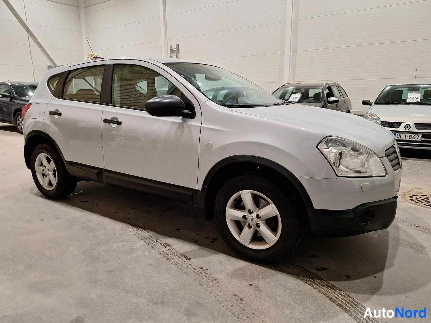Etuviisto, nokka oikealle: Hopeanharmaa Nissan Qashqai etukulmasta oikealta kuvattuna autotallissa, muiden autojen takana.