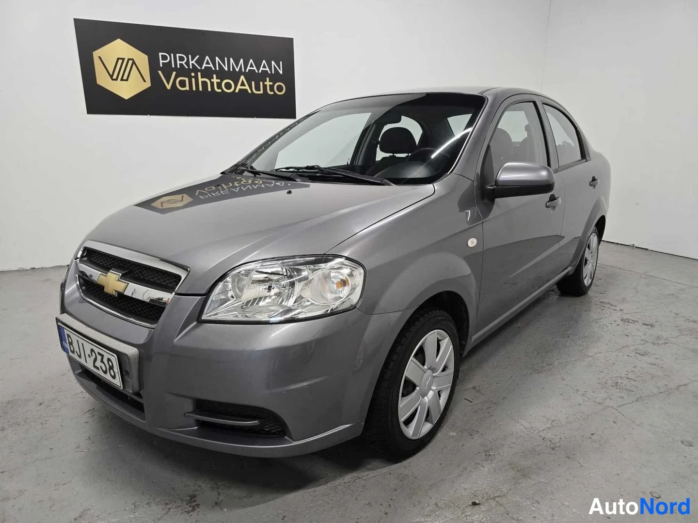 Etuviisto, nokka vasemmalle: Chevrolet Aveo -sedanin etukulma vasemmalta valokuvattuna harmaana sisätiloissa.