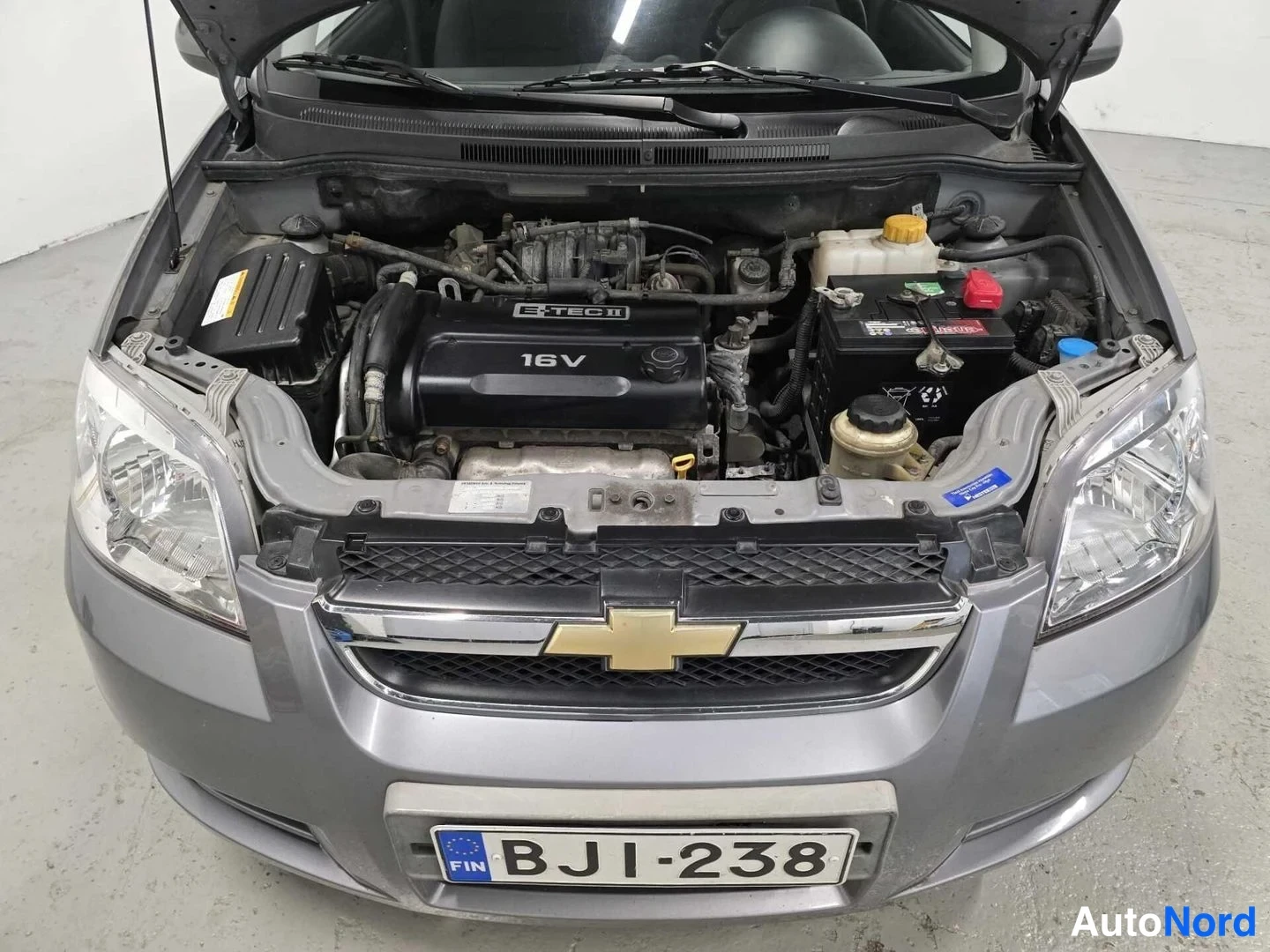 Edestä: Chevrolet Aven motorrum med öppen motorhuv, motorcover märkt 'F-TEC II 16V', kylarvätskebehållare och batteri syns, registreringsskylt.