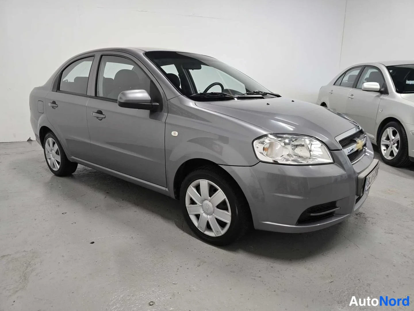 Etuviisto, nokka oikealle: Harmaa Chevrolet Aveo -sedanin etukulma oikealta vasemmalle valokuvattuna sisätiloissa.
