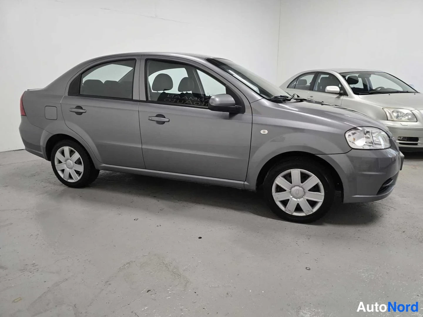 Etuviisto, nokka oikealle: Chevrolet Aveo -sedanin etukulma kuvattuna oikealta; vaaleanharmaa koripinta, etusäleikkö, ajovalot ja oikeanpuoleiset ovien kahvat ja vanteet näkyvät.