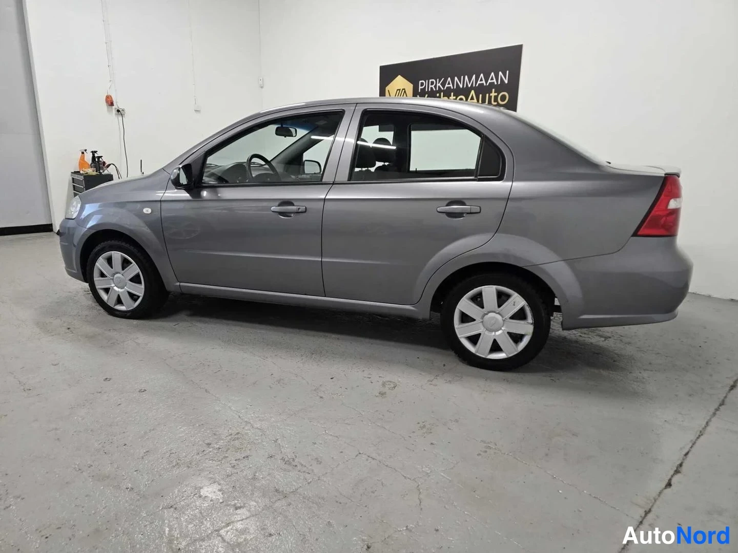 Takaviisto: Chevrolet Aveo -sedanin takakulma kuvattuna oikealta; vaaleanharmaa koripinta, punainen takavalo ja takapyörän hopeavanne näkyvät valkoisessa autotallissa.