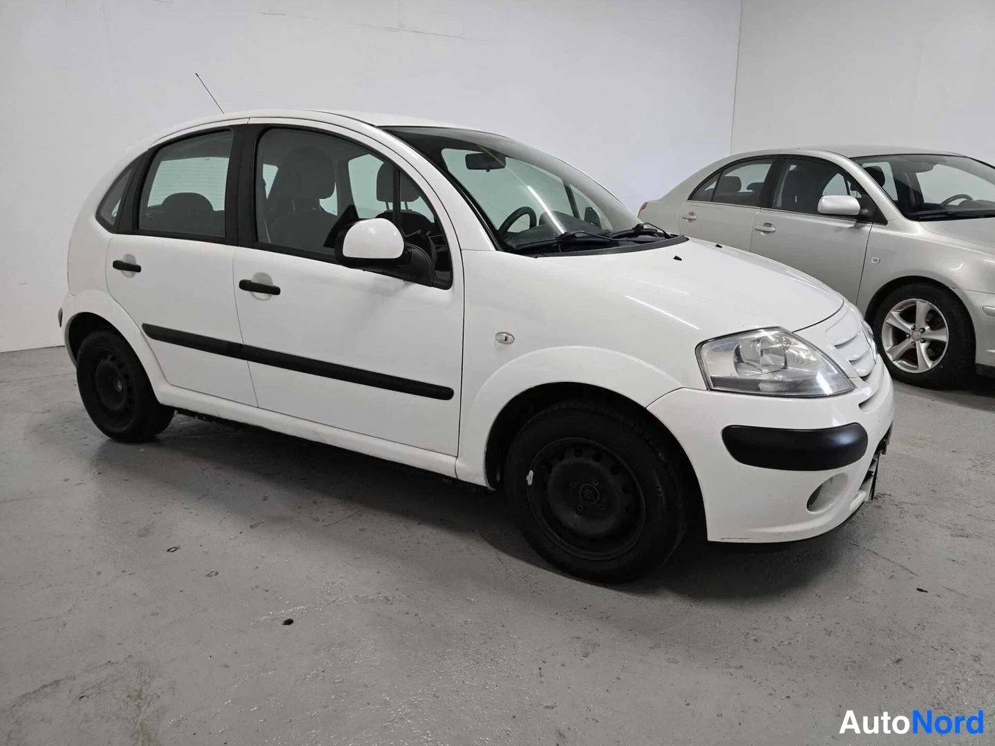 Etuviisto, nokka oikealle: Valkoisen Citroën C3:n etukulma vasemmalta kuvattuna autotallissa, toinen auto taustalla.