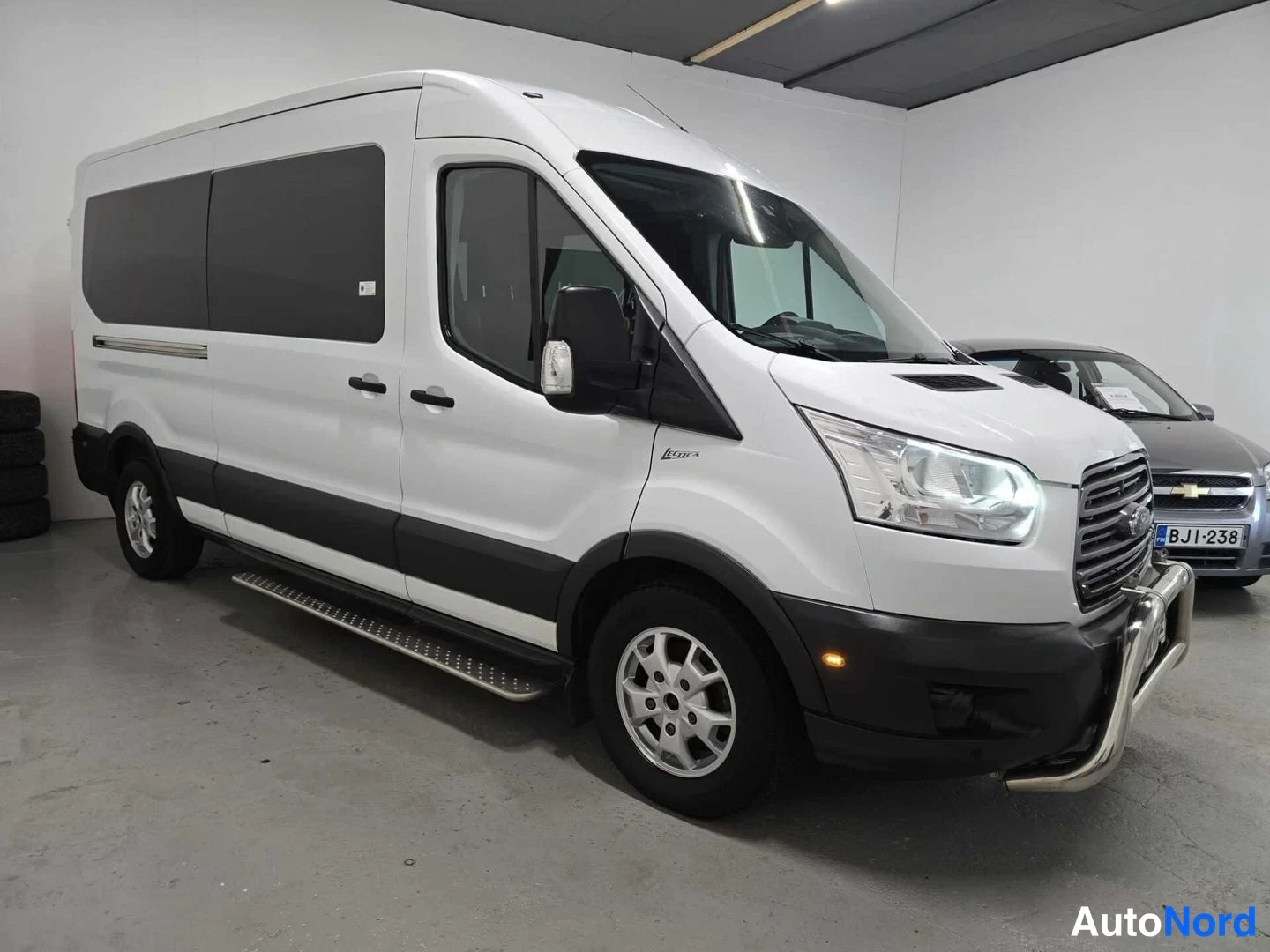 Etuviisto, nokka oikealle: Valkoinen Ford Transit -pakettiauto kulmasta edestä oikealta kuvattuna; etusäleikkö ja sivut näkyvät, vieressä toinen ajoneuvo.