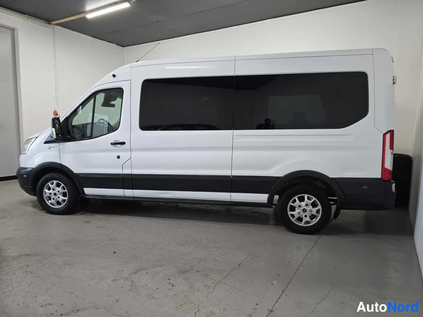 Sivulta: Valkoinen Ford Transit -pakettiauto sivulta kuvattuna; kuljettajan puoli, tummat ikkunat ja pyörät näkyvät.