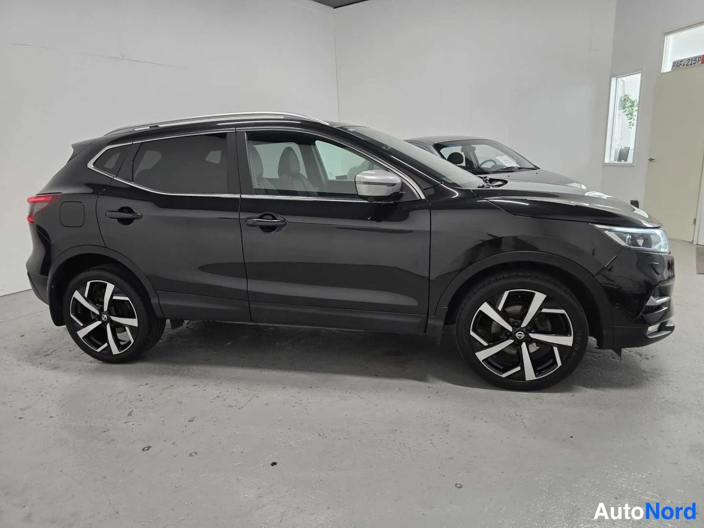 Etuviisto, nokka oikealle: Mustan Nissan Qashqai -maasturin etunurkkakuva oikealta, jossa näkyvät etupuskuri, ajovalot, kylki ja tummat kevytmetallivanteet sisätiloissa.