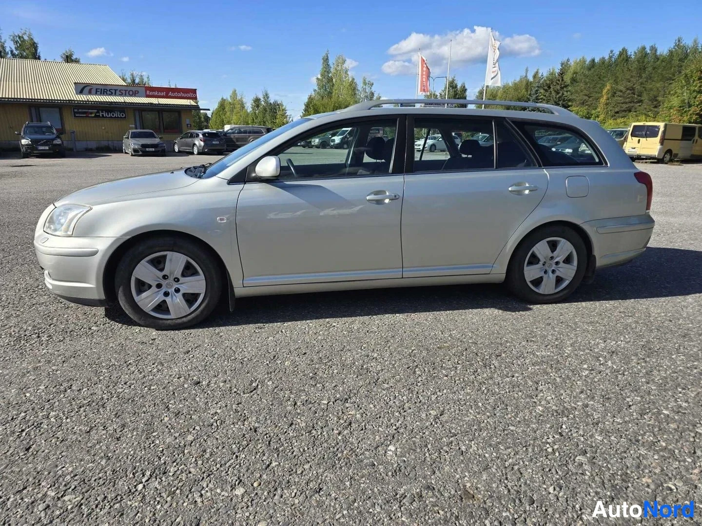 Sivulta: Toyota Avensis -farmari sivulta vasemmalta katsottuna, hopeanvärinen kori, kattokaiteet, ovenkahvat ja peilit näkyvissä.