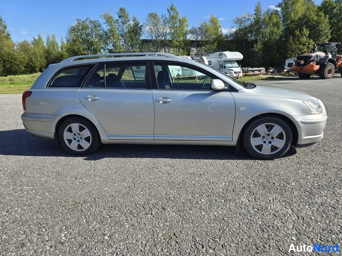 Sivulta: Toyota Avensis -farmari sivulta oikealta katsottuna, hopeanvärinen kori, kattokaiteet ja tummat ovenkahvat näkyvissä.