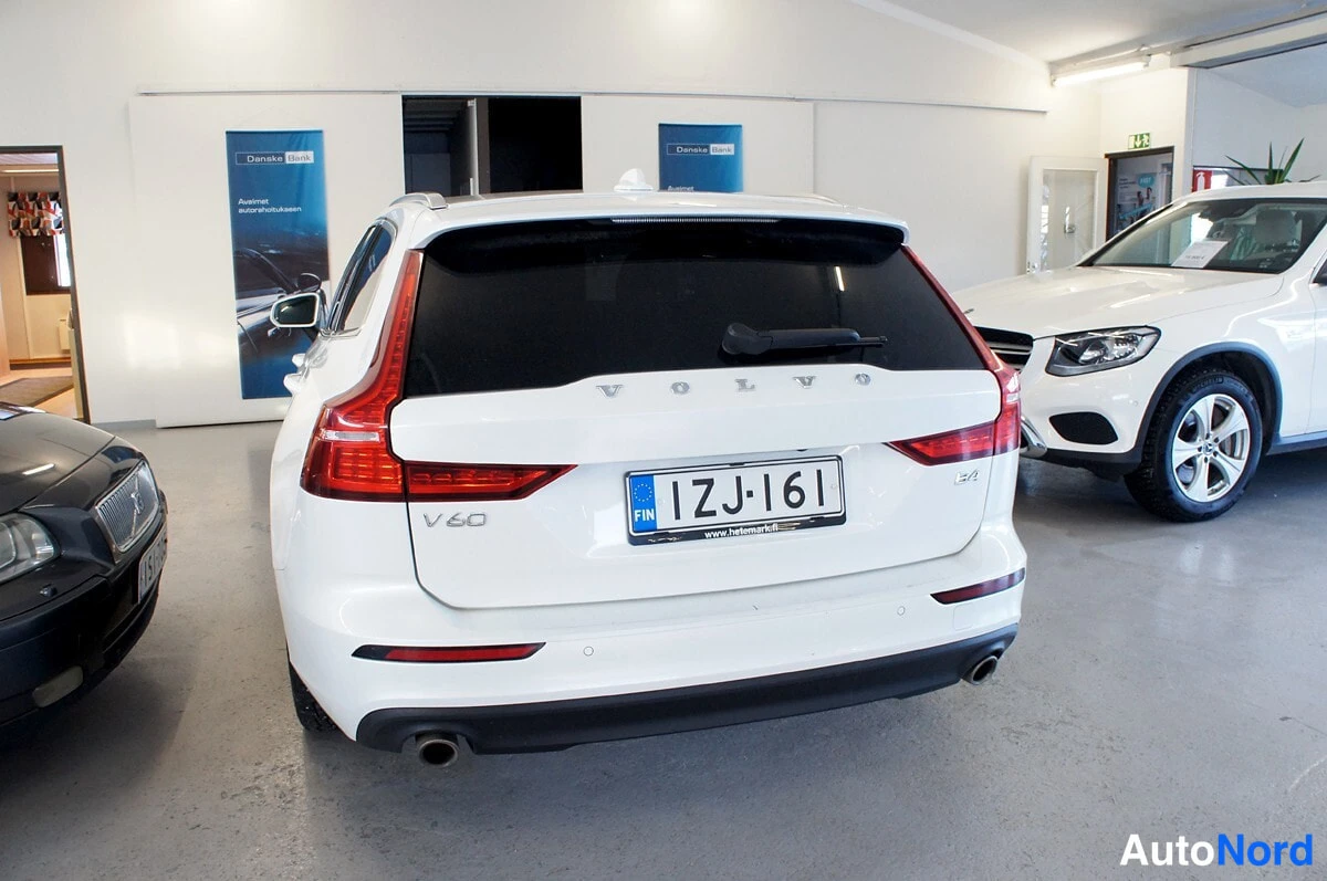 Takaa: Valkoisen farmariauton suora takaosan näkymä sisätiloissa: VOLVO- ja V60-tekstit takaluukussa, punaiset takavalot ja rekisterikilpi 1ZJ-161.