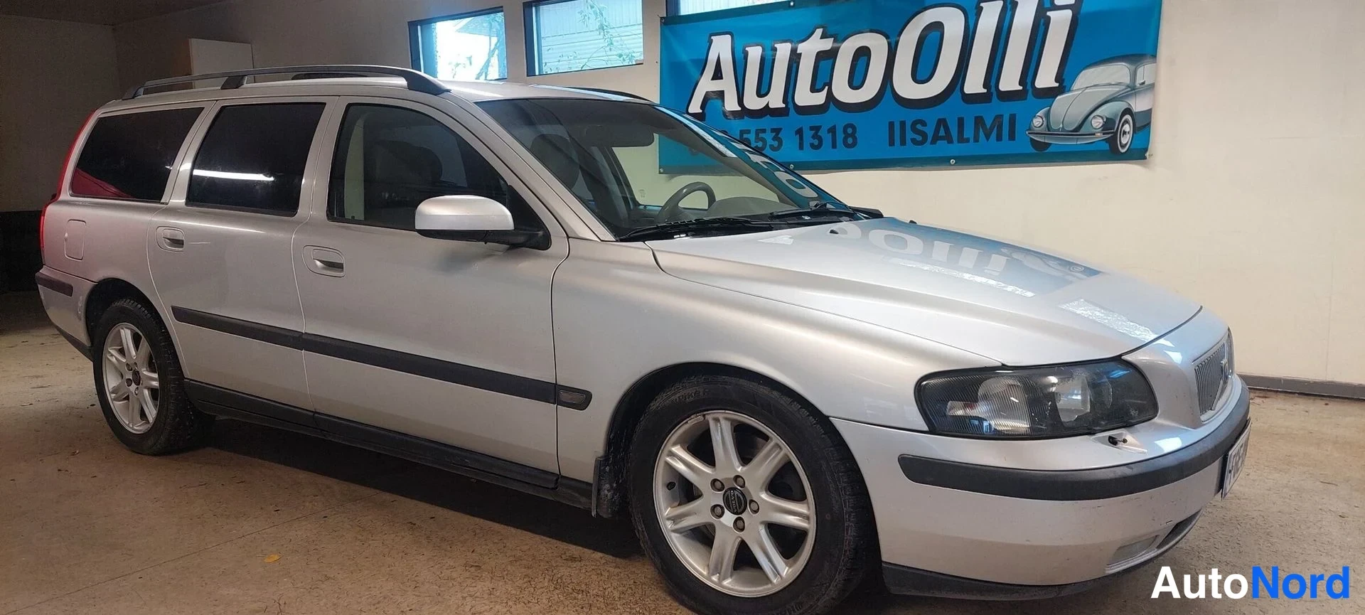 Etuviisto, nokka oikealle: Hopeinen Volvo V70 -farmari vasemmasta etukulmasta, keula oikealla, AutoOlli-mainoskyltti taustalla.