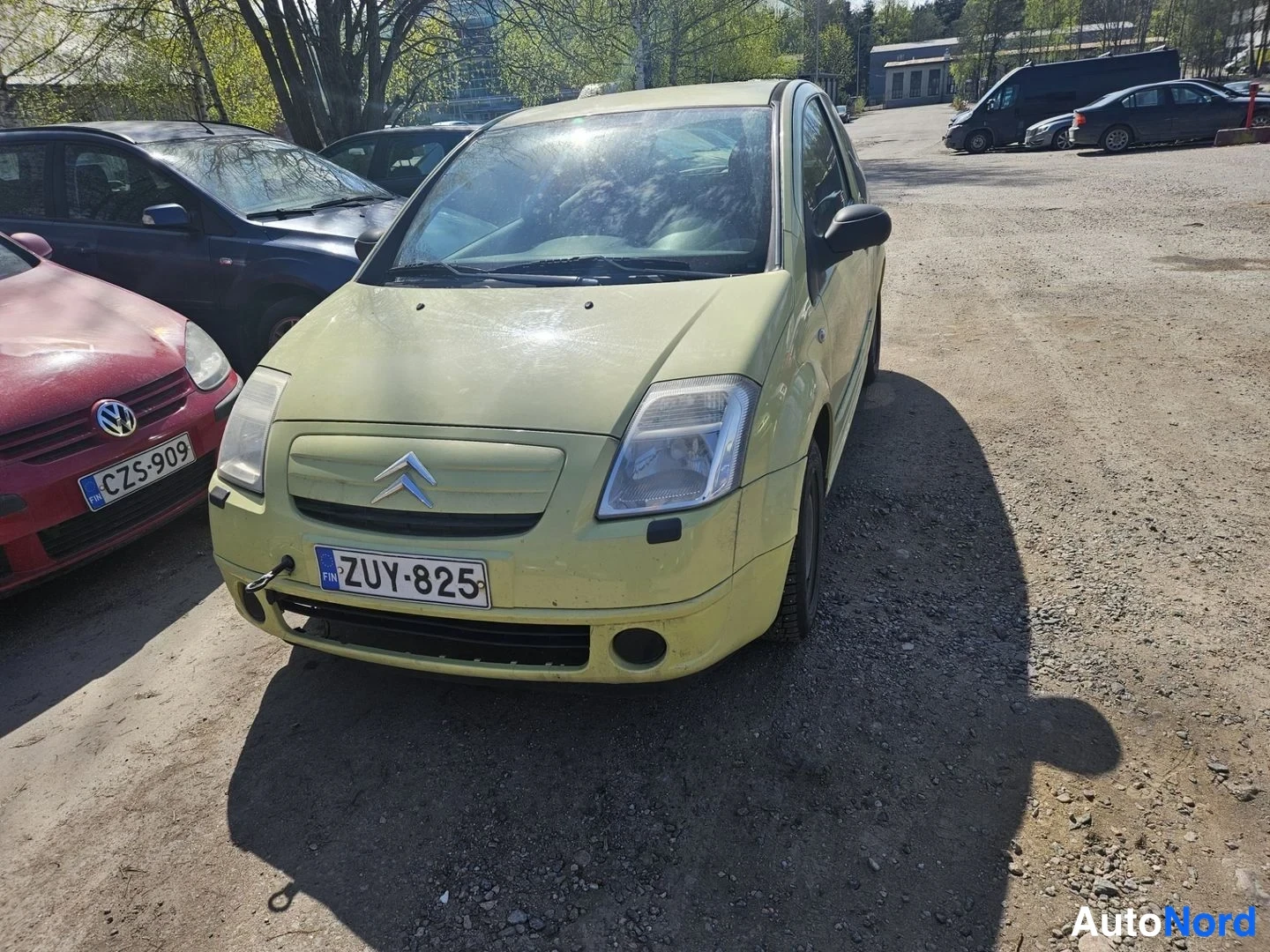 Etuviisto, nokka vasemmalle: Citroën C2 etuosalta vasemmasta kulmasta, etulasi heijastaa aurinkoa, rekisterikilpi, ympärillä muita pysäköityjä autoja.