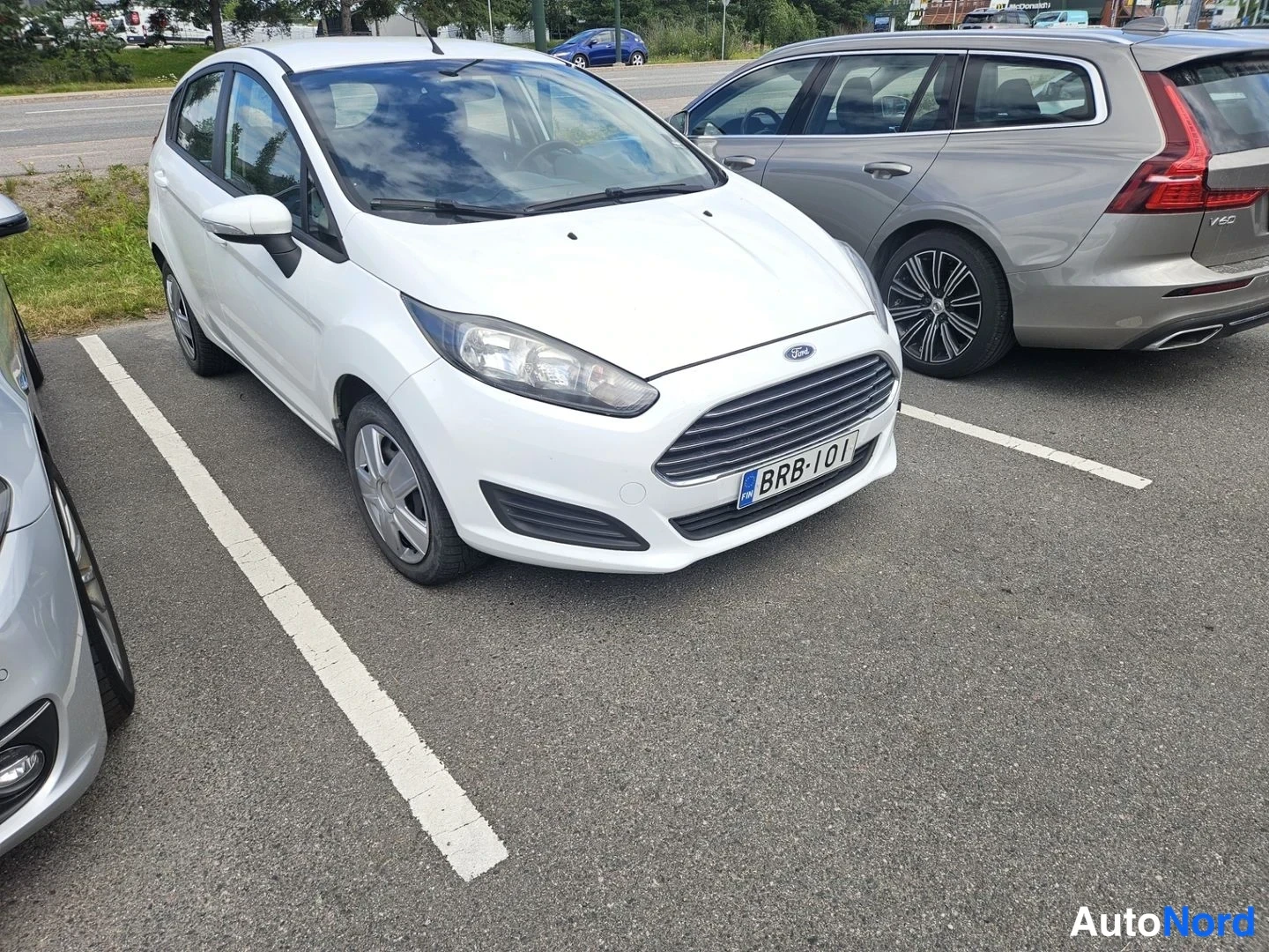 Etuviisto, nokka oikealle: Valkoinen Ford Fiesta etualalla pysäköintipaikalla, etunäkymä oikealta kolmasosan kulmasta.