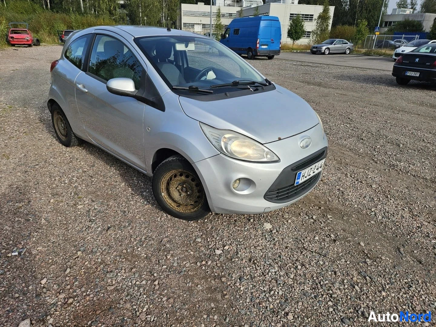 Etuviisto, nokka oikealle: Hopeanvärinen Ford Ka kulmauksesta edestä oikealta päin kuvattuna sorapohjaisella parkkipaikalla, muita ajoneuvoja ja rakennuksia taustalla.
