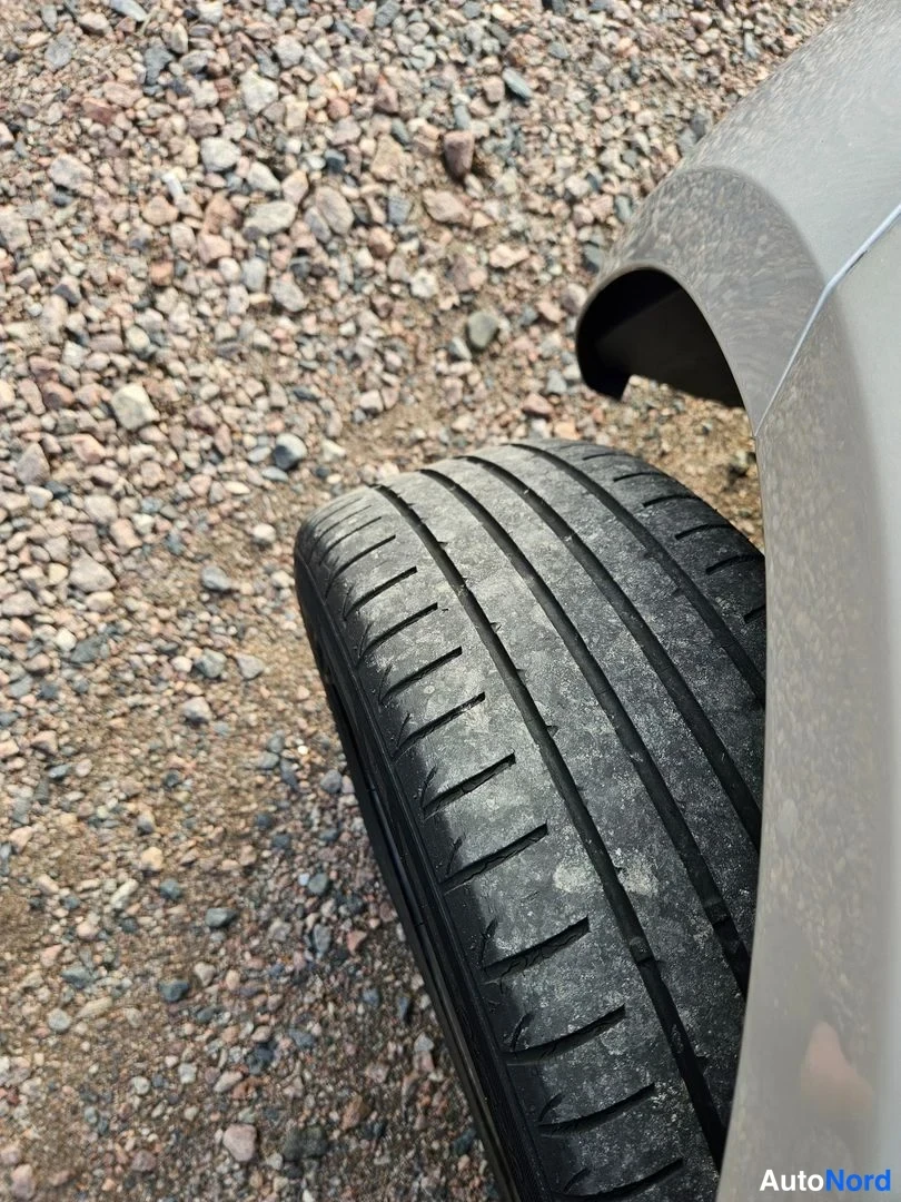 Yksityiskohta: Auton renkaan ja etukolmanneksen sivusta lähi­kuva sora­li­vannolla: keski­kar­reas­sa kulunut kesä­rengas ja kevyt­metalli­vanne, kori­paneeli vaalean­harmaa.