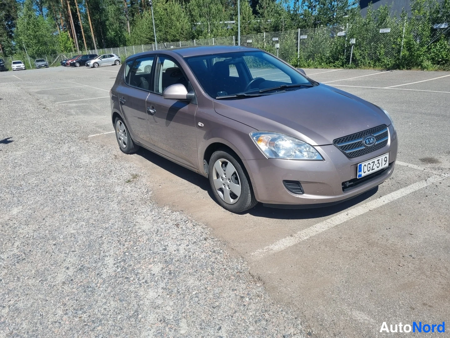 Etuviisto, nokka oikealle: Kia Ceed -henkilöauto tuhkanruskeassa värissä kolmessa neljäsosasta edestä oikealle kuvattuna parkkipaikalla.