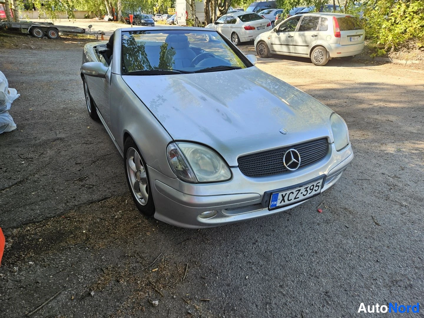 Etuviisto, nokka oikealle: Hopeanvärinen avokattoinen Mercedes SLK urheiluauto etukulmasta oikealta, rekisteritunnus.