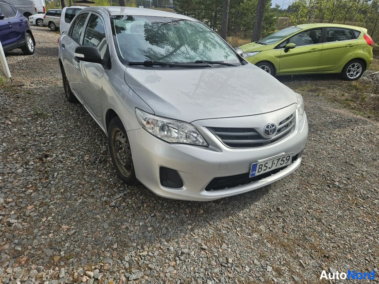 Etuviisto, nokka oikealle: Hopeanvärinen Toyota Corolla -henkilöauto kuvattu edestä oikealta, rekisteritunnus.