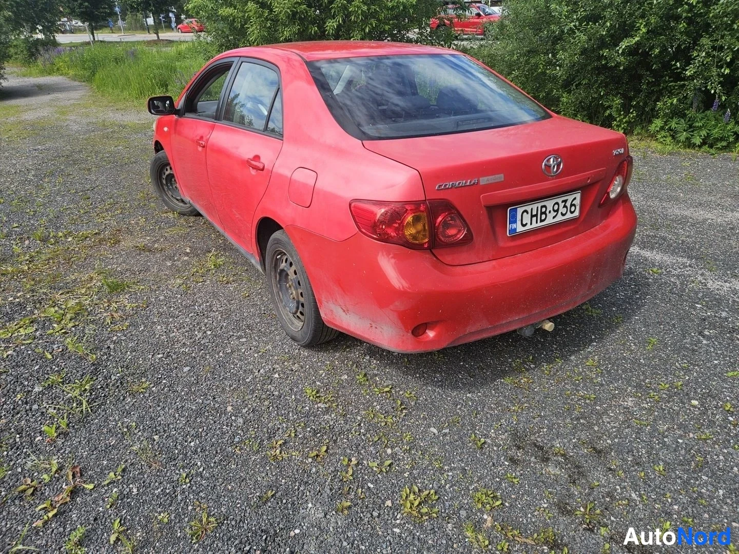 Takaviisto: Punaisen Corolla-sedanin takakulma oikealta: Corolla-merkintä ja Toyota-emblem takaluukussa, rekisteritunnus, pinttyneitä naarmuja takapuskurissa.