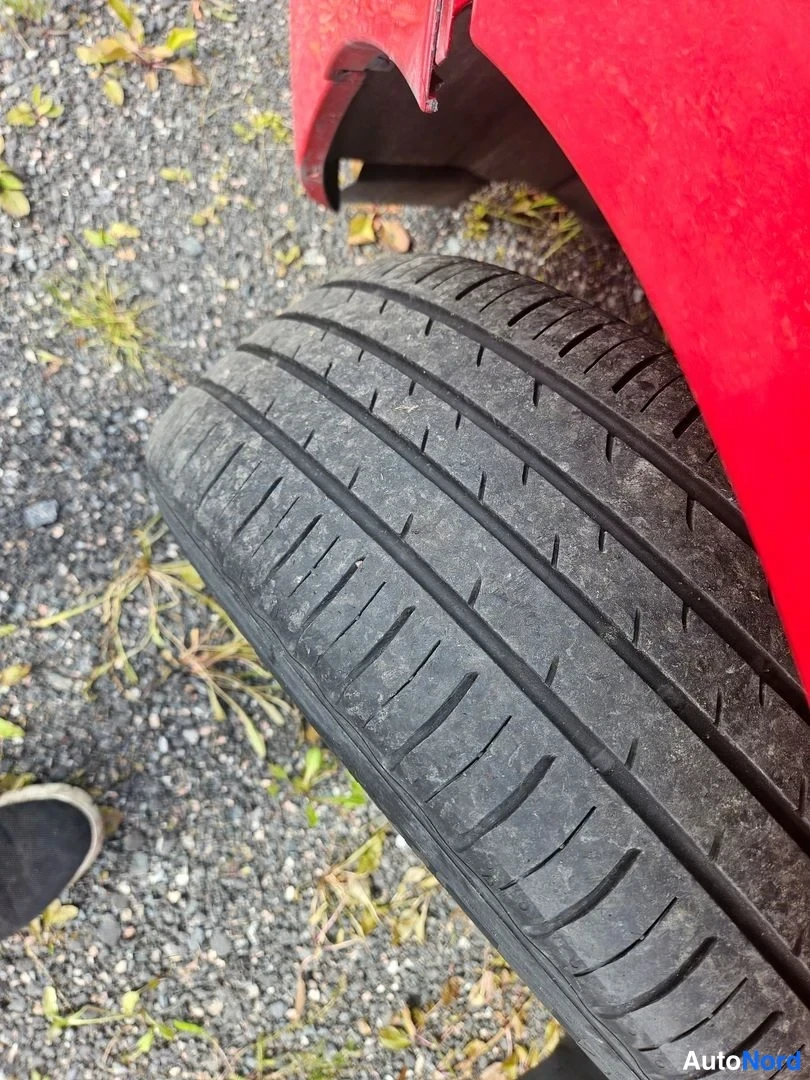 Yksityiskohta: Lähikuva auton mustasta renkaasta: kulutuspinnan urat ja kulumisjäljet näkyvät tarkasti, punainen runkopaneeli yläreunassa ja sora-asfaltti taustalla.