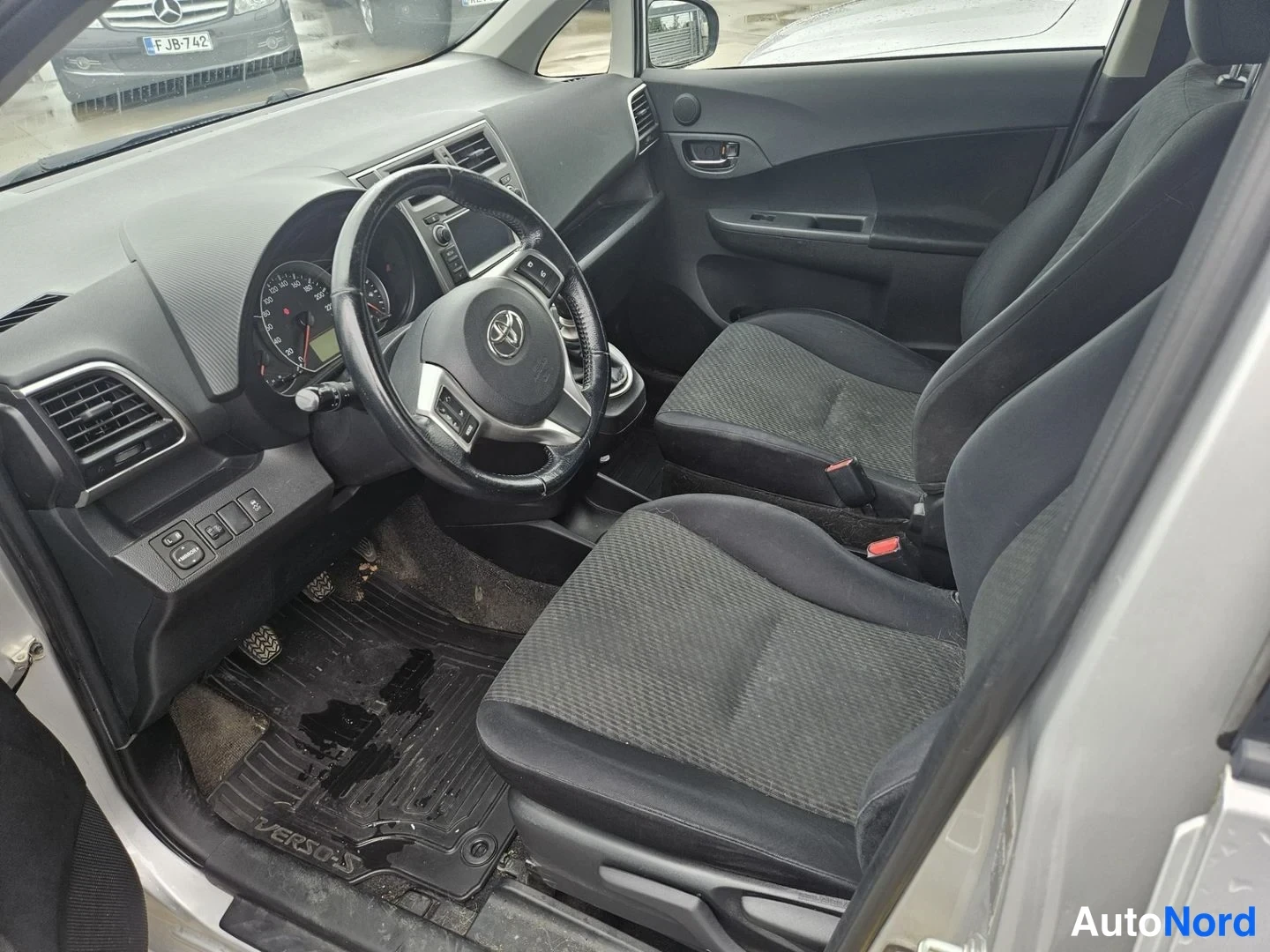 Muu: Auton etuistuimien ja kojelaudan sisäkuva kuljettajan oven puolelta: Toyota-ohjauspyörä, tummat kangasistuimet, manuaalivaihteisto ja lattiassa 'VERSO-S'-teksti