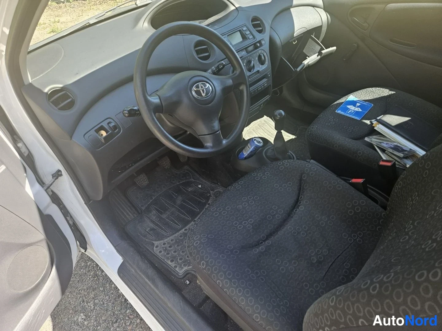 Ohjaamo: The driver's side interior of the car, featuring a black fabric seat with a circular pattern, an automatic gear selector lever, a steering wheel with the Toyota