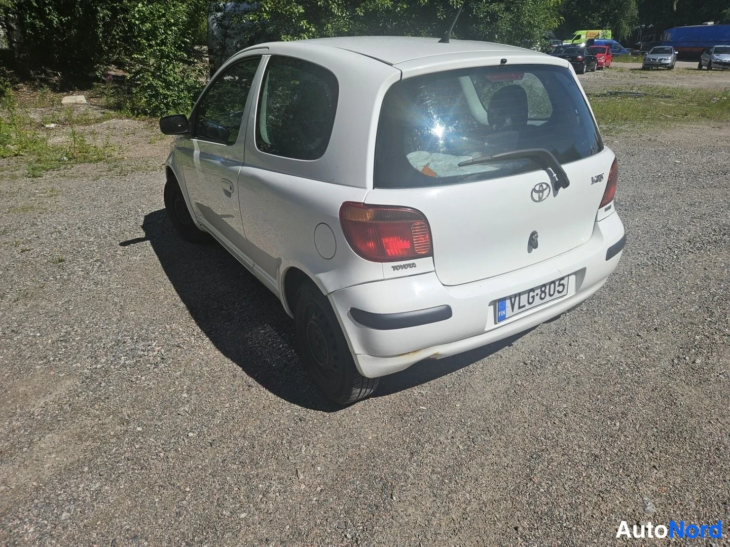 Takaviisto: Valkoinen Toyota Yaris -henkilöauto takaa kolmesta neljäsosasta kuvattuna sorapäällysteisellä parkkialueella.