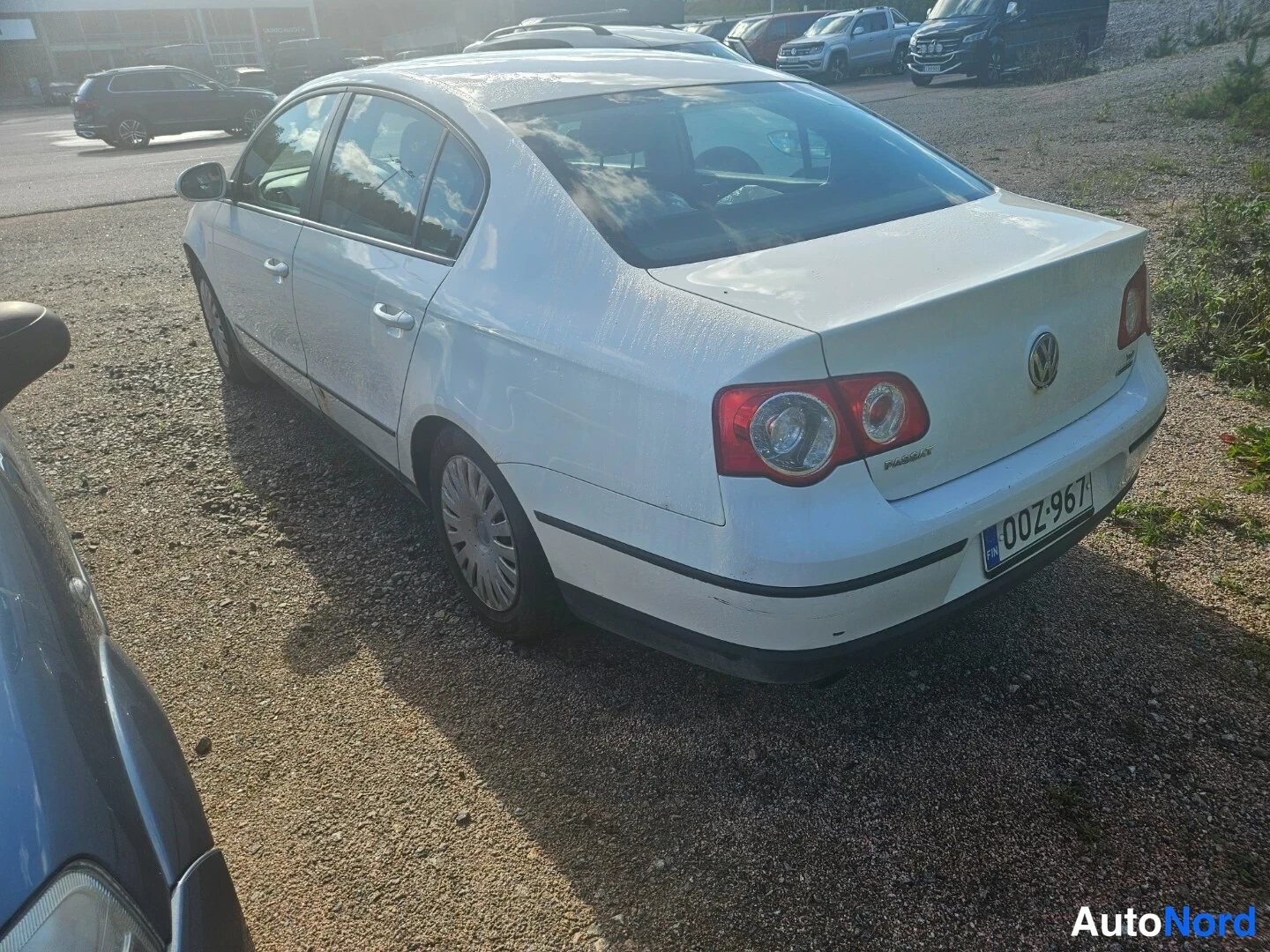 Takaviisto: Valkoinen Volkswagen Passat -henkilöauto takaosalta ja vasemmalta sivulta kuvattuna soraparkissa.