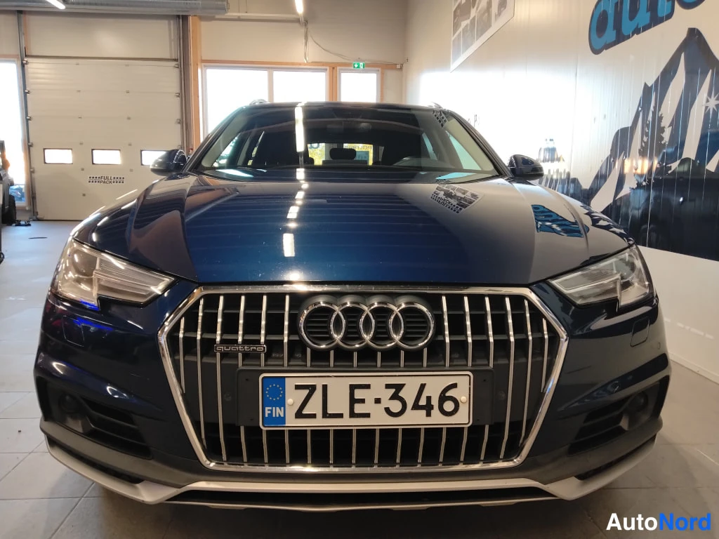 Edestä: Tummansininen käyttöauto suoraan edestä sisätiloissa; etusäleikkö Audi-merkeillä ja rekisterikilpi näkyvät.