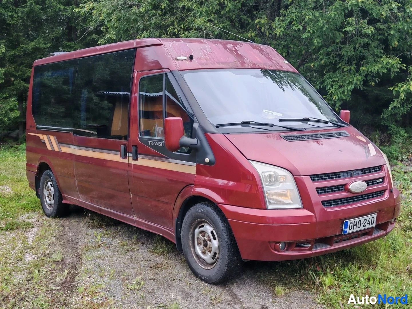 Etuviisto, nokka oikealle: Punaruskea Ford Transit -bussi kolmannelta sivulta edestä, keula oikealla, rekisterikilpi.