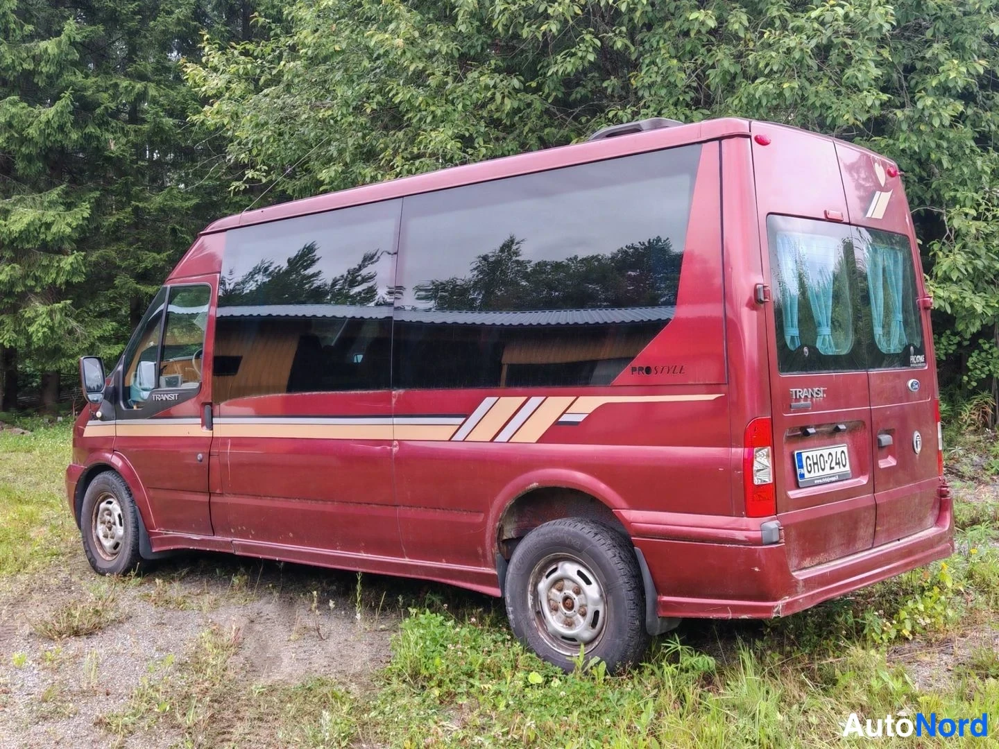 Takaviisto: Punaruskean Ford Transit -bussin vasen sivu ja takaosa, takaikkunoissa siniset verhot, rekisterikilpi.