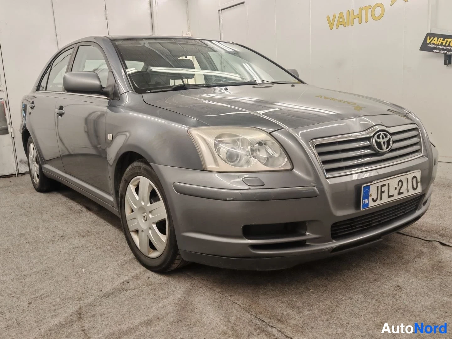 Etuviisto, nokka oikealle: Tummanharmaa Toyota Avensis -sedan etuokolikuva vasemmalta katsoen, etusäleikkö ja rekisterikilpi näkyvissä.