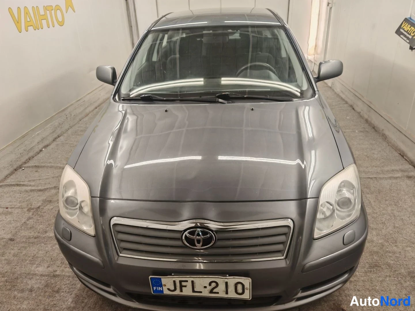 Edestä: Tummanharmaa Toyota Avensis -sedan suora etunäkymä sisätiloissa, keulakoriste ja rekisterikilpi keskitettynä.