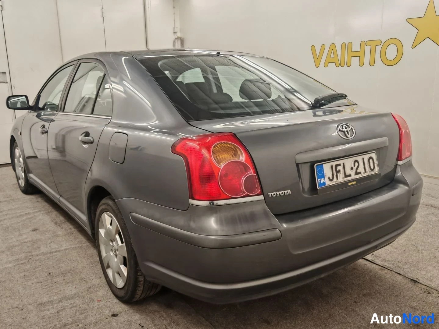 Takaviisto: Harmaa Toyota Avensis takaviistosta vasemmalta: takaosa ja vasen kylki näkyvissä, rekisterikilpi, hopeavat pyöränvanteet.